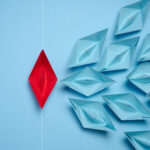 A group of blue paper boats and one red sailing against the current, concept of independent personality, breaking free from influence, strong leadership